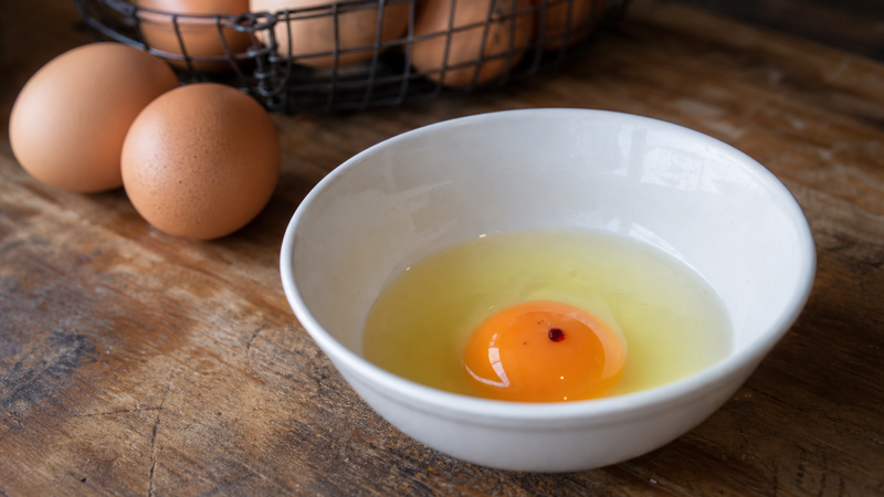 Cracked backyard chicken egg in a white bowl showing a small red blood spot on the yolk